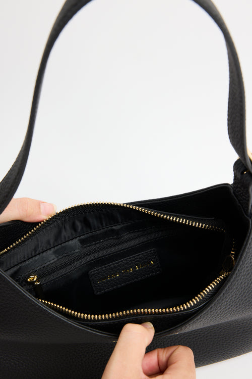 Close-up of a black pebbled leather handbag interior with a gold zipper, shown with two hands holding the opening. A small black interior label is visible.