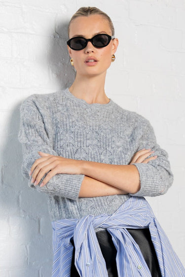 Person wearing a gray sweater, light blue shirt, black skirt, and black shoes against a white wall.