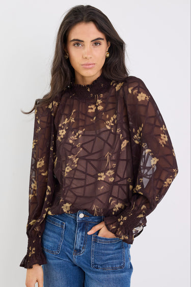 Woman wearing a sheer brown floral blouse with blue jeans against a white background