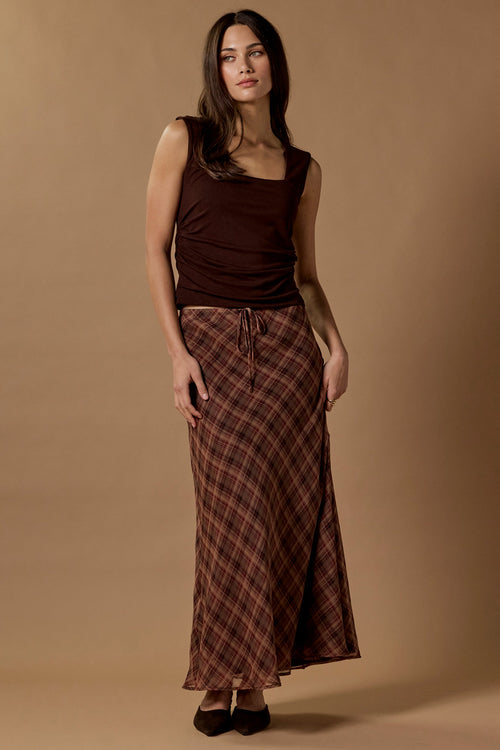 A woman modeling a dark brown sleeveless top and a long brown and red plaid skirt against a solid beige studio backdrop.