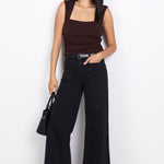 Woman wearing a brown top and black wide-leg pants holding a black handbag on a white background