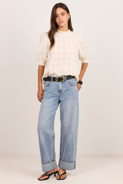 A woman modeling a cream-colored textured puff-sleeve top and light-wash wide-leg blue jeans with a black belt, standing against a plain off-white studio background.
