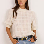 A woman modeling a cream-colored textured puff-sleeve blouse with a subtle plaid pattern, tucked into light-wash blue jeans with a black belt, standing against a plain off-white wall.