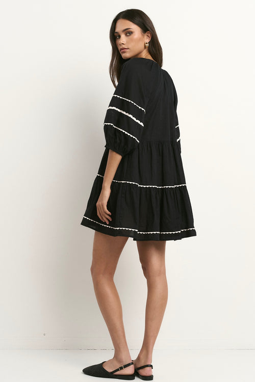 Woman wearing a black mini dress with tiered skirt, white scalloped trim, and puff sleeves.