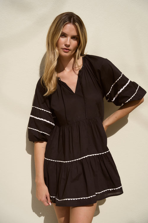 Woman wearing a black mini dress with tiered skirt, white scalloped trim, and puff sleeves.