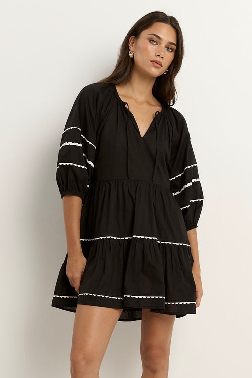 Woman wearing a black mini dress with tiered skirt, white scalloped trim, and puff sleeves.