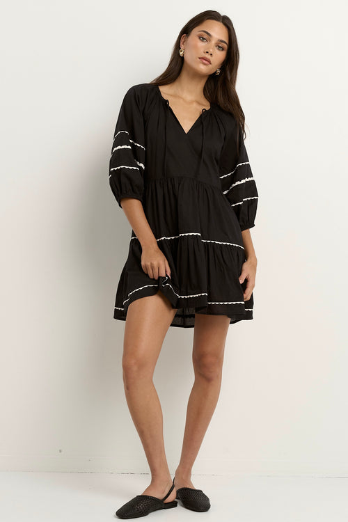 Woman wearing a black mini dress with tiered skirt, white scalloped trim, and puff sleeves.