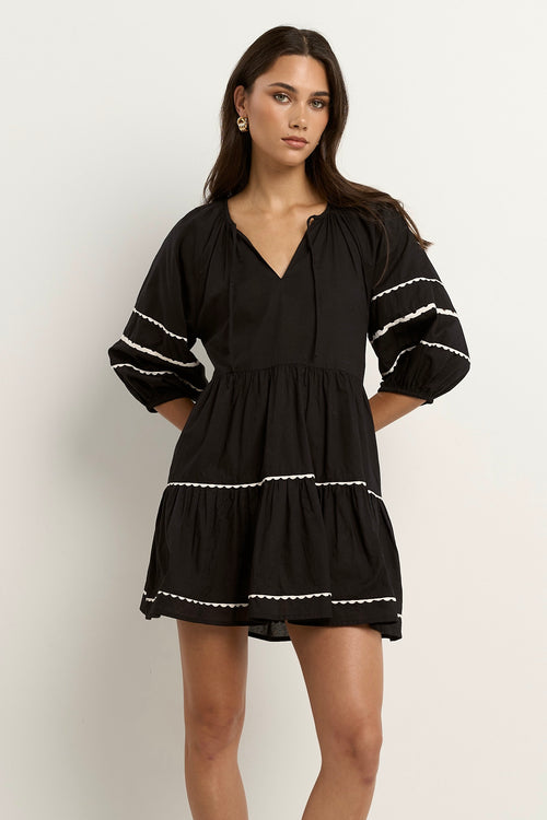 Woman wearing a black mini dress with tiered skirt, white scalloped trim, and puff sleeves.