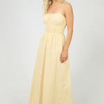 Woman wears a lemon yellow maxi dress with a smocked bodice and thin spaghetti straps. The dress flows gently from the waist, styled with strappy heels for a light, airy feel.