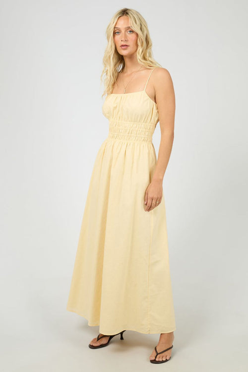 Woman wears a lemon yellow maxi dress with a smocked bodice and thin spaghetti straps. The dress flows gently from the waist, styled with strappy heels for a light, airy feel.