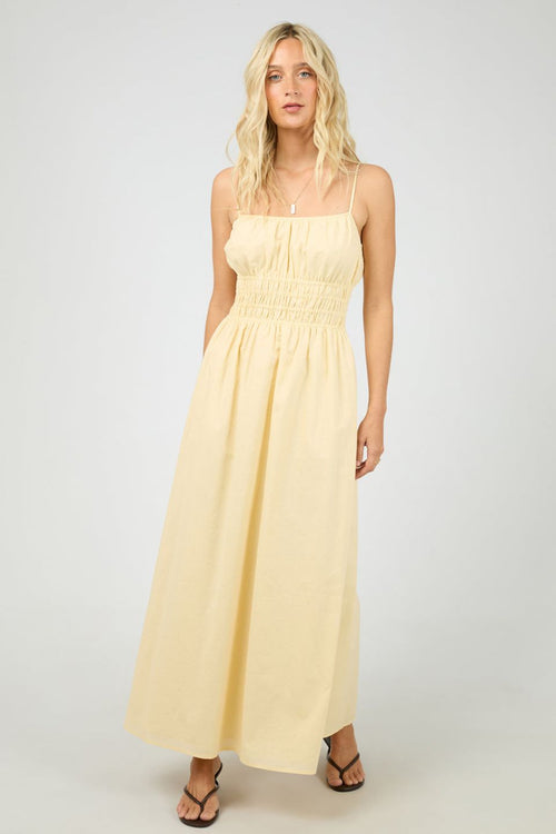 Woman wears a lemon yellow maxi dress with a smocked bodice and thin spaghetti straps. The dress flows gently from the waist, styled with strappy heels for a light, airy feel.
