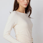 A woman wearing a cream-colored long-sleeve top with ruching at the waist, paired with blue jeans, photographed against a light gray studio background.