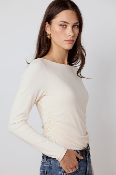 A woman wearing a cream-colored long-sleeve top with ruching at the waist, paired with blue jeans, photographed against a light gray studio background.