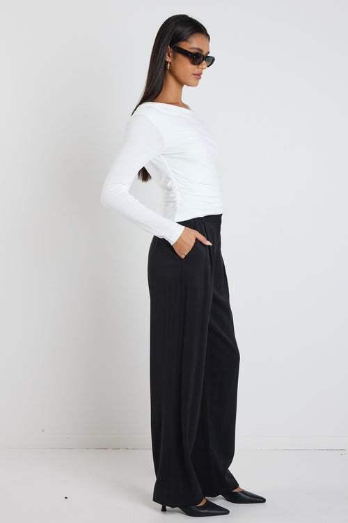 A woman wearing an ivory long-sleeved top with a wide bateau neckline and subtle ruched side detailing, styled with black pants.