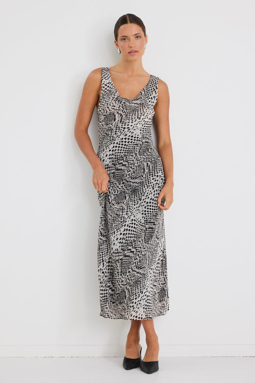 Woman wearing a sleeveless, bias-cut midi dress with a charcoal snake print, styled with black heels against a white wall.