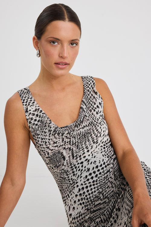 Woman wearing a sleeveless, bias-cut midi dress with a charcoal snake print, styled with black heels against a white wall.