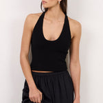 A woman modeling a black sleeveless halter crop top and a matching black high-waisted maxi skirt with a side slit, styled with black sunglasses, black sandals, and a small black handbag against a plain white studio background.