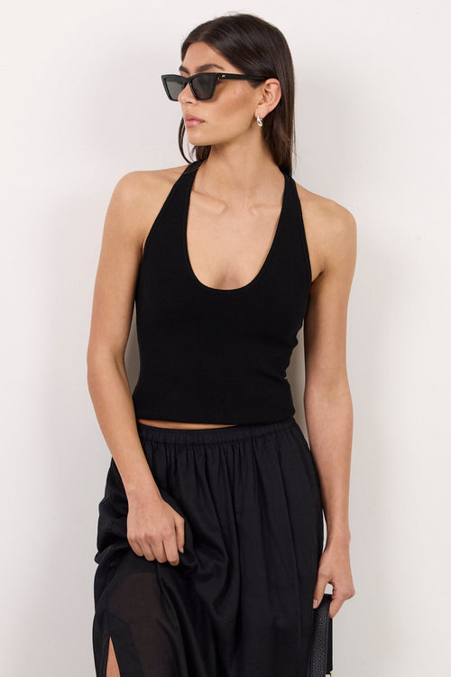 A woman modeling a black sleeveless halter crop top and a matching black high-waisted maxi skirt with a side slit, styled with black sunglasses, black sandals, and a small black handbag against a plain white studio background.