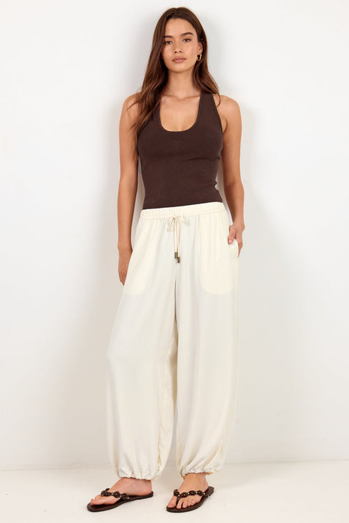 A woman modeling a dark brown ribbed tank top and cream-colored wide-leg drawstring pants with black sandals, standing against a plain white studio background.