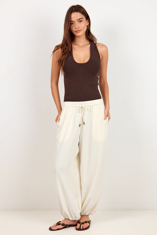 A woman modeling a dark brown ribbed tank top and cream-colored wide-leg drawstring pants, paired with dark brown sandals, against a plain white studio background.