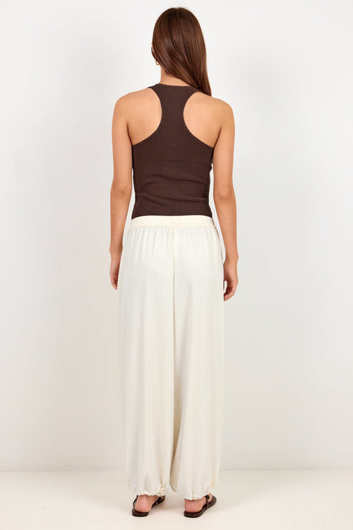 A woman shown from the back wearing a dark brown racerback tank top and white wide-leg pants, standing against a plain white studio background.