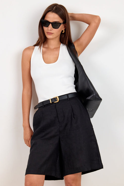 A woman modeling a white ribbed tank top, black pleated shorts with a black belt, black sunglasses, and carrying a black shoulder bag against a plain white background.
