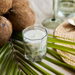 A small hexagonal glass candle holder with a white candle and a printed green palm leaf design, styled on green palm leaves with coconuts and a glass goblet in the background.