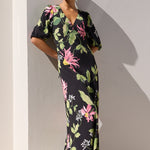 A woman wearing a black maxi dress with a vibrant pink and green tropical floral print stands barefoot by a swimming pool against a white wall.