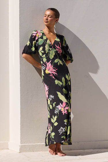A woman wearing a black maxi dress with a vibrant pink and green tropical floral print stands barefoot by a swimming pool against a white wall.