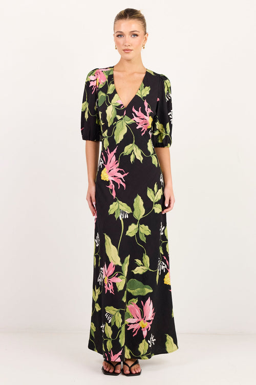 A woman modeling a black maxi dress with a vibrant pink and green tropical floral print, standing against a plain white studio background.