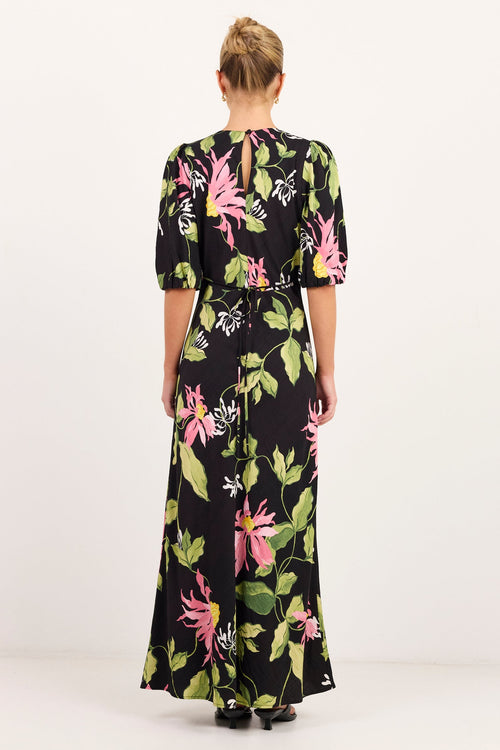 A woman viewed from behind wearing a black maxi dress with a vibrant green, pink, and white tropical floral print, standing against a plain white studio background.