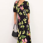 A woman modeling a black maxi dress with a vibrant pink and green floral print, styled with a black handbag and sunglasses against a plain white studio background.