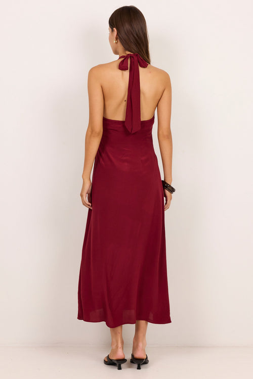 A woman viewed from the back wearing a sleeveless halter-neck maxi dress in deep burgundy, paired with black pointed-toe flats, set against a plain off-white studio background.
col_pattern