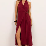 A woman modeling a sleeveless halter-neck midi dress in deep burgundy with a wrap-style front and high side slit, standing against a plain off-white studio background.