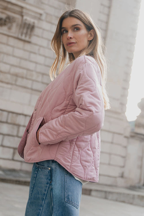 Woman wearing a pink quilted jacket standing in front of a stone building.