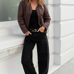 Woman standing against a white building wearing a brown jacket and black pants.