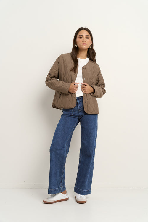 Woman wearing an oversized quilted mocha colored jacket with button front, styled with blue jeans and gold hoop earrings.