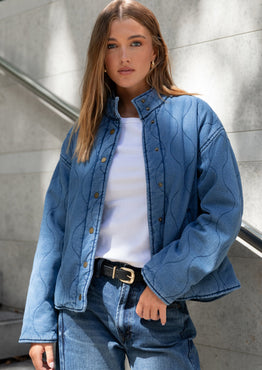 Woman wearing a blue denim jacket and jeans standing against a gray wall.
