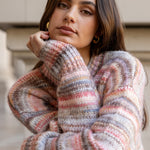 Woman wearing a colorful striped sweater and blue jeans standing against a neutral background