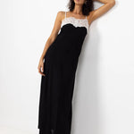 A woman modeling a black maxi dress with a white lace-trimmed spaghetti strap bodice, leaning against a white wall.