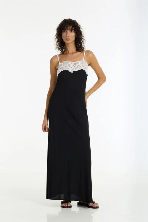 A woman modeling a long black slip dress with thin straps and a white lace bust detail, standing against a plain white studio background.
