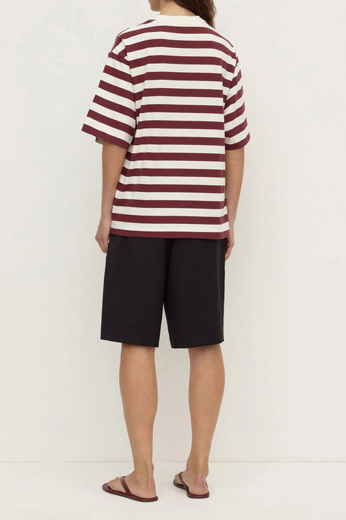 A person shown from the back wearing a short-sleeve striped t-shirt with burgundy and off-white horizontal stripes, paired with black shorts and brown sandals, against a plain off-white studio background.