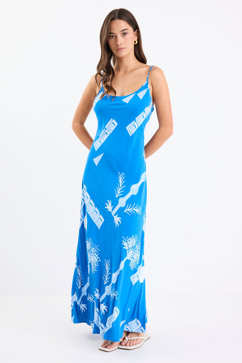 Woman wearing a bright blue strappy bias-cut slip midi dress with white palm and geometric print, relaxed fit, and fluid drape.
