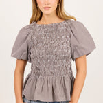 A woman modeling a short-sleeve smocked blouse with a subtle gray and white gingham pattern, paired with light-wash blue jeans, against a plain off-white studio background.