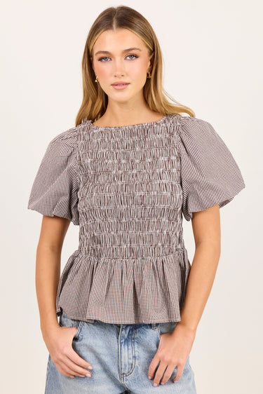 A woman modeling a short-sleeve smocked blouse with a subtle gray and white gingham pattern, paired with light-wash blue jeans, against a plain off-white studio background.