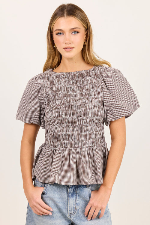 A woman modeling a short-sleeve smocked blouse with a subtle gray and white gingham pattern, paired with light-wash blue jeans, against a plain off-white studio background.