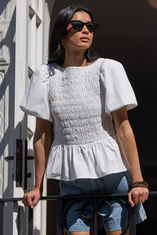 A woman wearing a white smocked puff-sleeve peplum top and denim shorts stands on a balcony in front of white architectural doors, wearing black sunglasses.