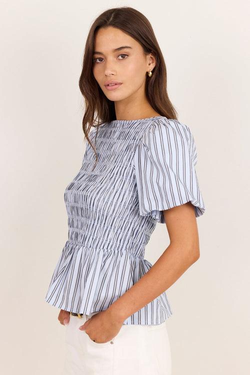 A woman modeling a blue and white vertically striped short-sleeve peplum top with a smocked bodice, paired with white pants, against a plain off-white background.