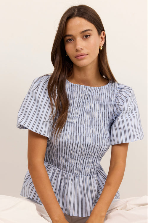 A woman wearing a blue and white vertically striped smocked blouse with puff sleeves, paired with white pants, sitting against a plain off-white background.
