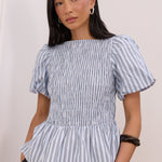 A woman modeling a blue and white vertically striped peplum blouse with puff sleeves, paired with black trousers, in an elegant indoor setting with white walls and molding.